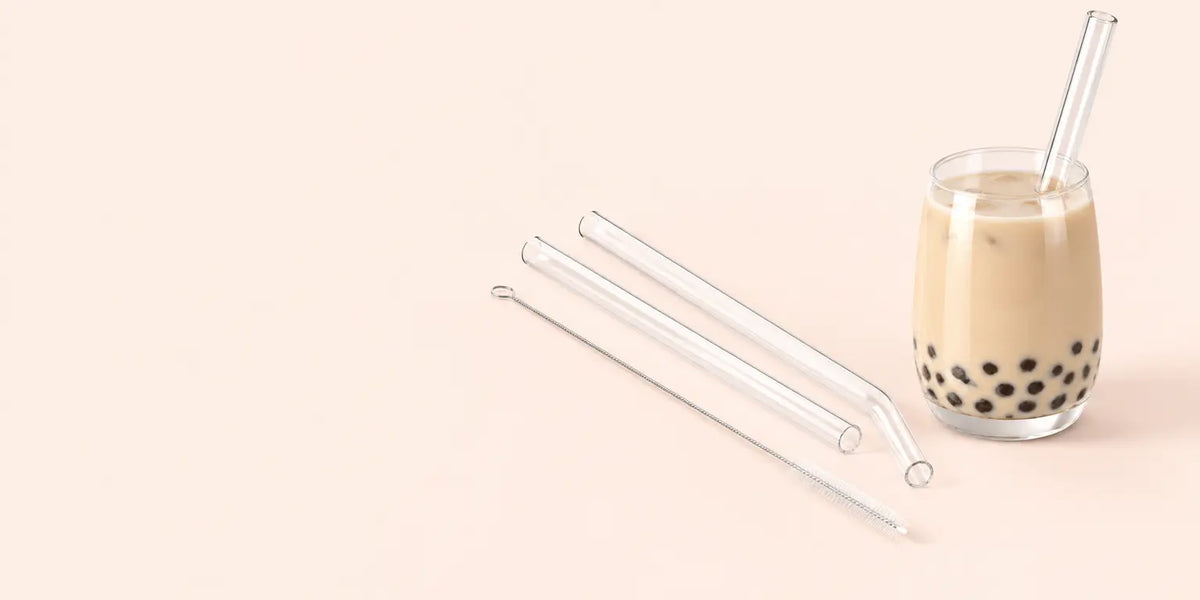 A studio photograph of exactly three clear borosilicate glass straws and their narrow cleaning brush
